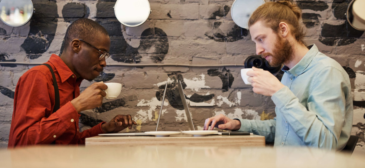 Side view portrait of two contemporary men drinking coffee and using laptops sitting at opposite sides of table in cafe, copy space